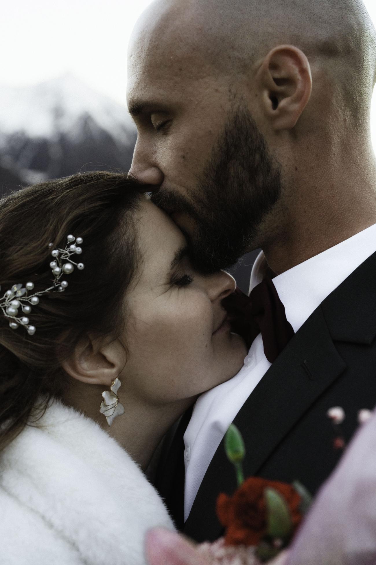 Couple de marié venant de la Rochelle pour se marier à saint lary de soulan, à l'hotel Christiana. Le marié embrasse avec beaucoup d'émotion le front de la mariée. En arrière plan, nous pouvons deviner les hautes-pyrénées enneigées. Cette photo est très forte émotionnellement et très élégante. 